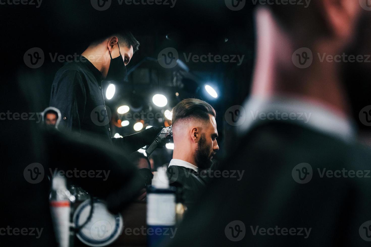 jovem barbudo sentado e cortando o cabelo na barbearia por cara com máscara protetora preta foto