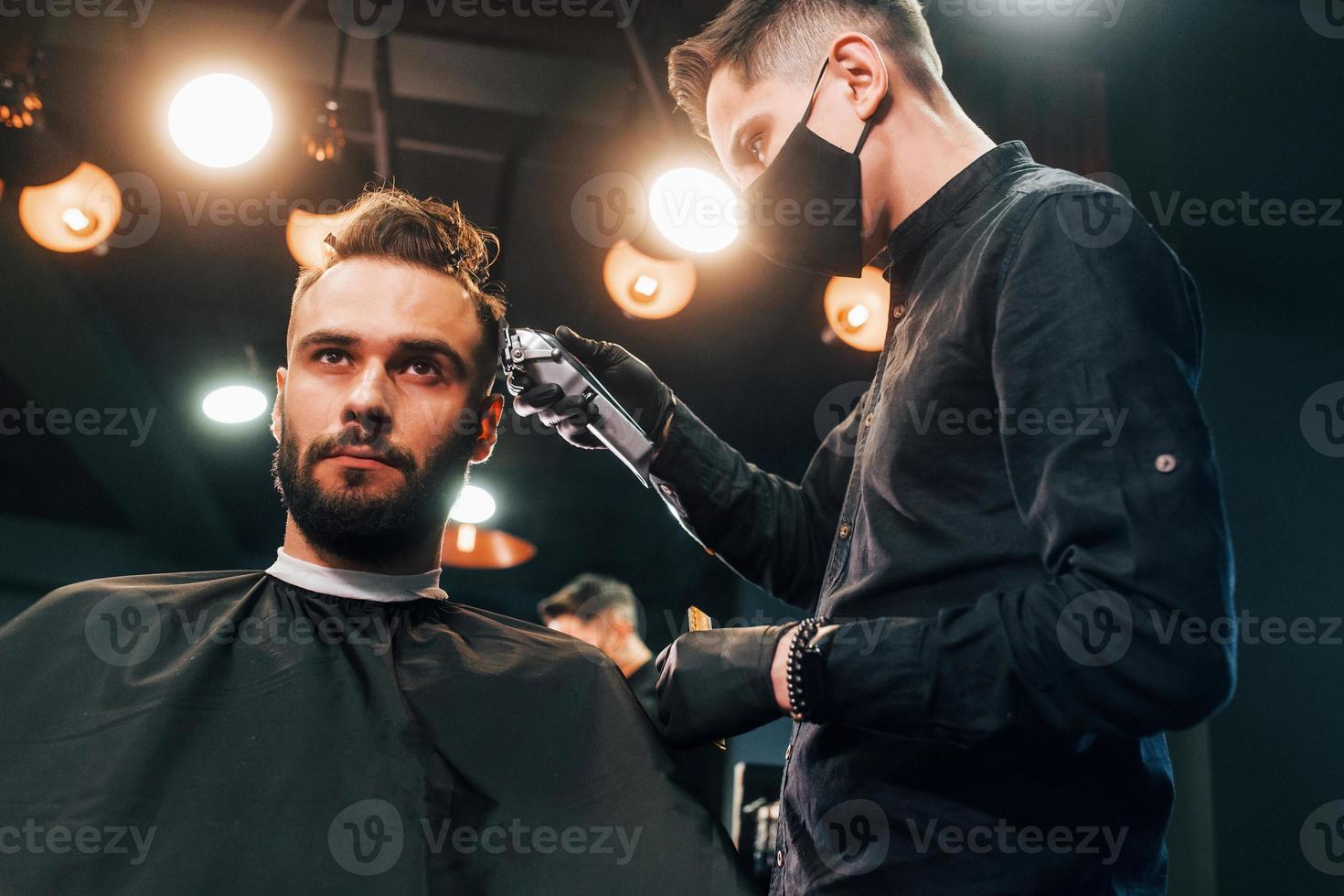 vista frontal do jovem barbudo sentado e cortando o cabelo na barbearia por cara com máscara protetora preta foto