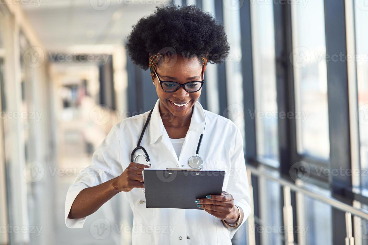 jovem médica afro-americana de uniforme branco com estetoscópio e bloco de notas em pé no corredor foto