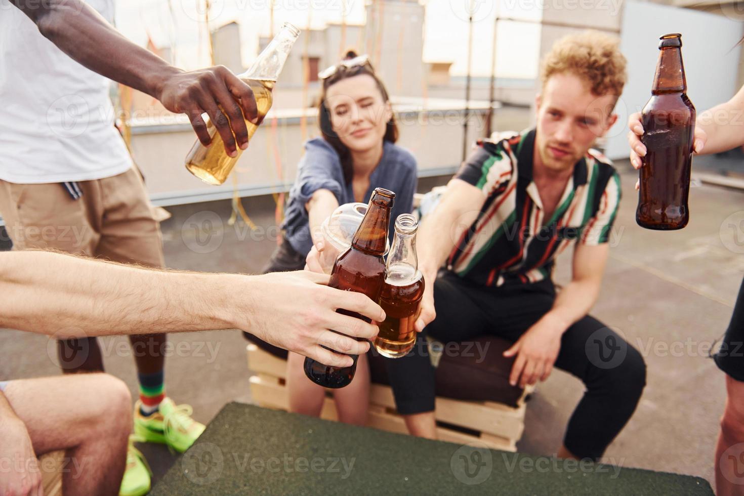 fazendo elogios por garrafas com cerveja. grupo de jovens em roupas casuais faz uma festa no telhado juntos durante o dia foto