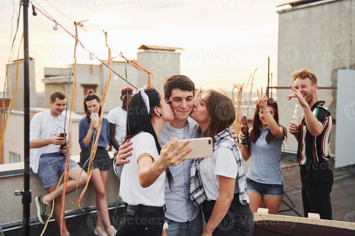 duas meninas beijando um homem. tirando selfie. grupo de jovens em roupas casuais faz uma festa no telhado juntos durante o dia foto