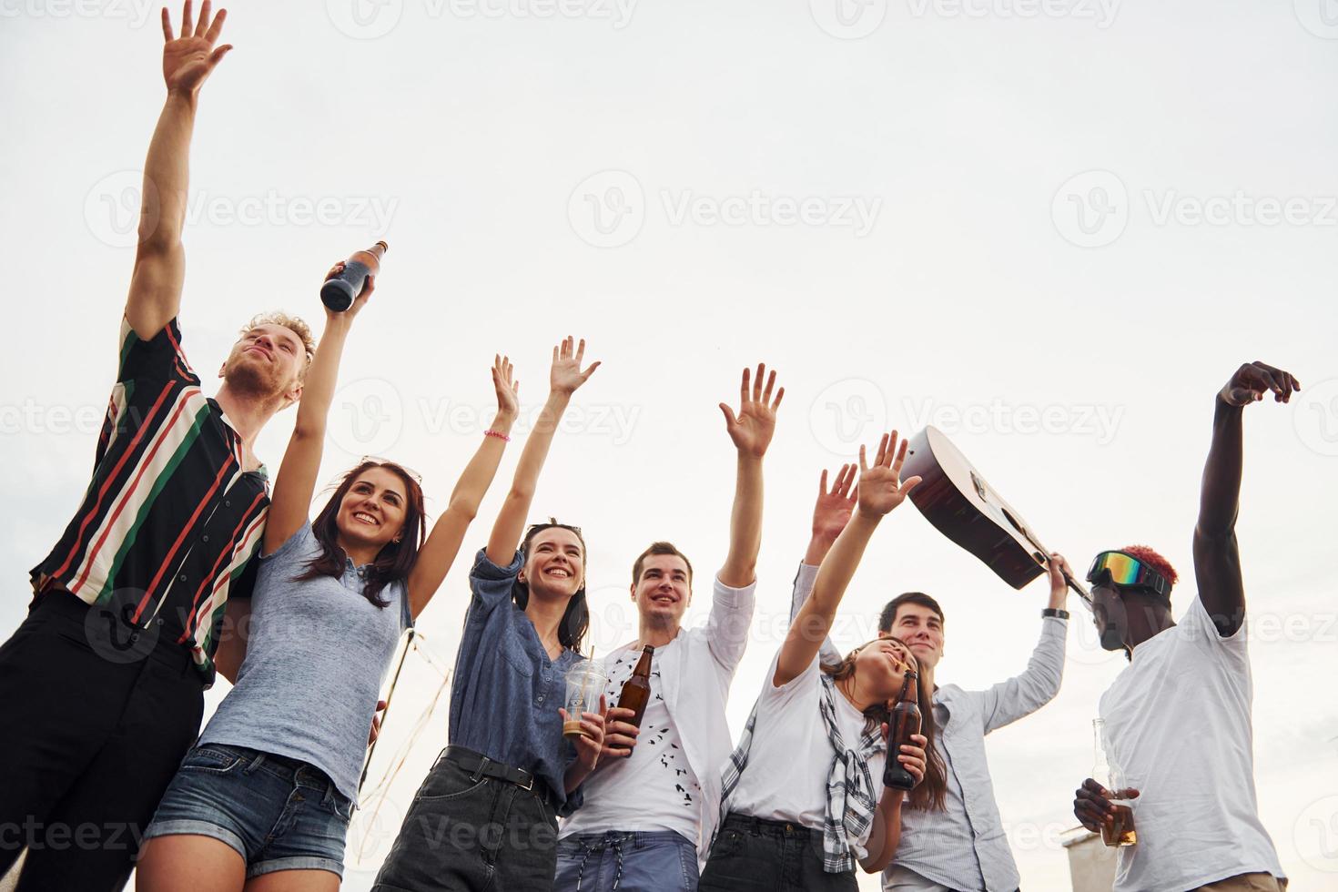 de pé alto e olhando para o céu nublado. grupo de jovens em roupas casuais faz uma festa no telhado juntos durante o dia foto