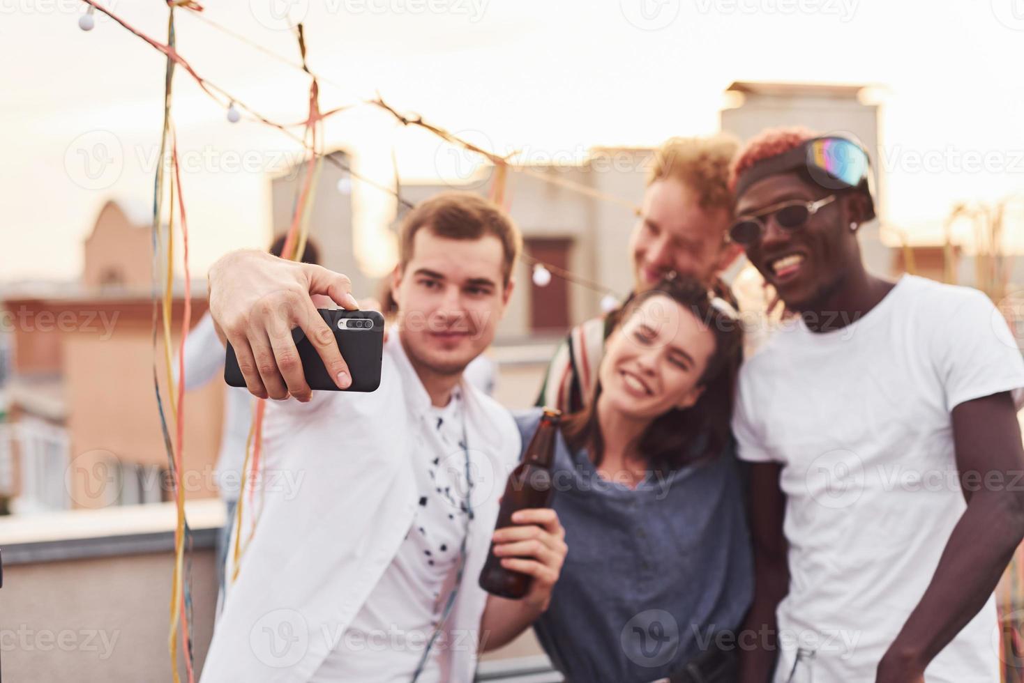 de pé juntos e tirando selfie. grupo de jovens em roupas casuais faz uma festa no telhado juntos durante o dia foto