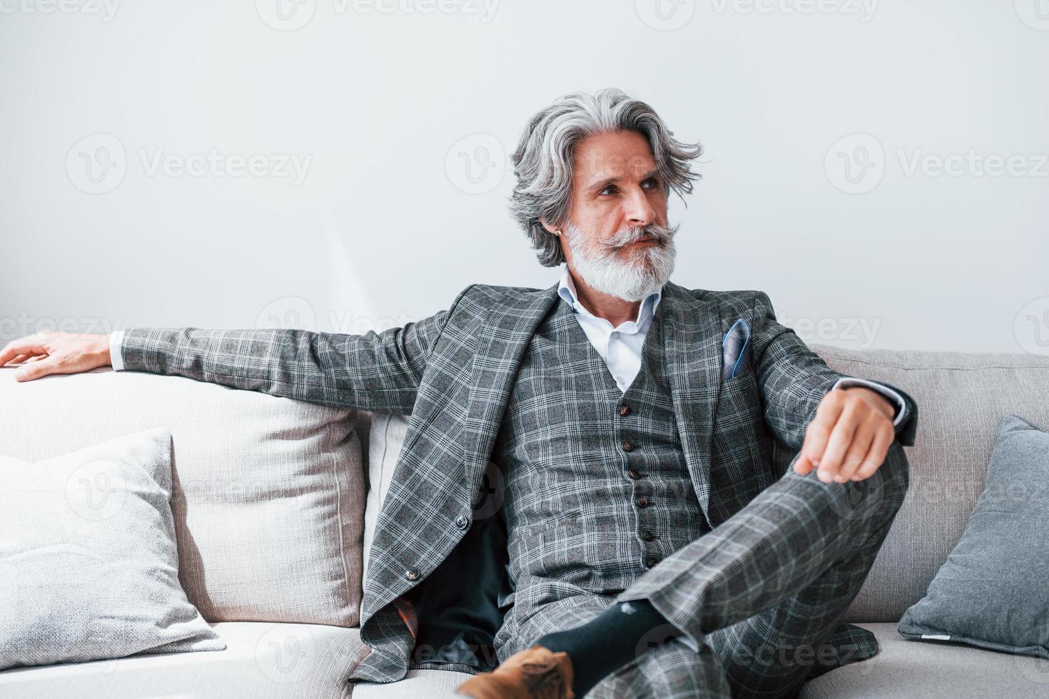 senta-se no sofá confortável em roupas formais. homem moderno elegante sênior com cabelos grisalhos e barba dentro de casa foto