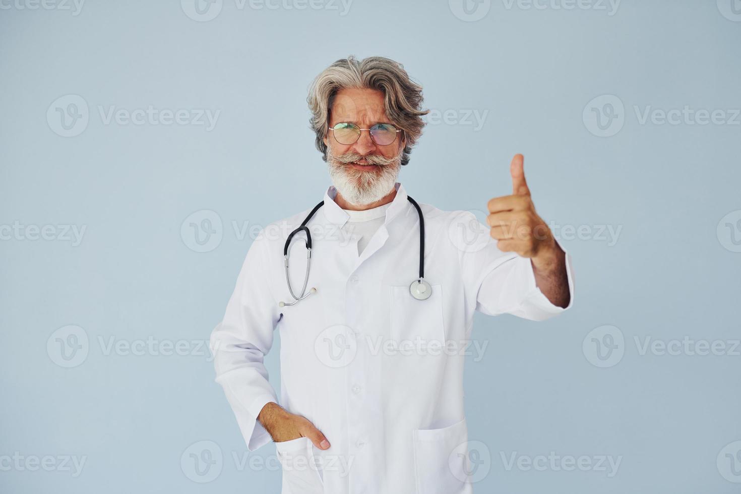 médico de jaleco branco contra a parede. homem moderno elegante sênior com cabelos grisalhos e barba dentro de casa foto