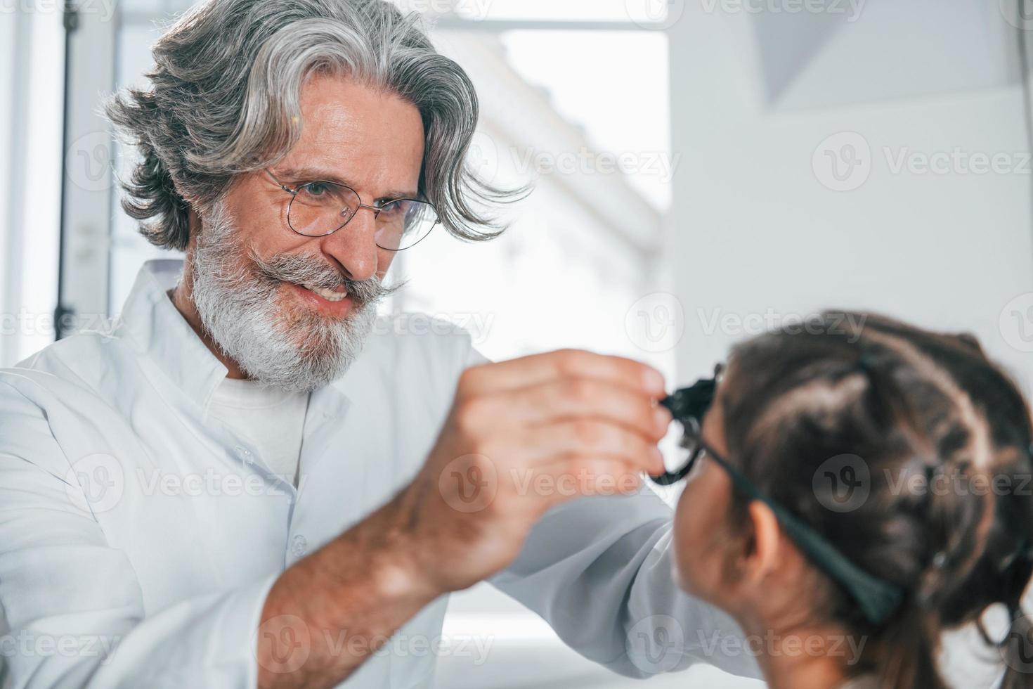 oftalmologista masculino sênior com cabelos grisalhos e barba em jaleco branco está dentro de casa na clínica testando a visão de uma garotinha foto