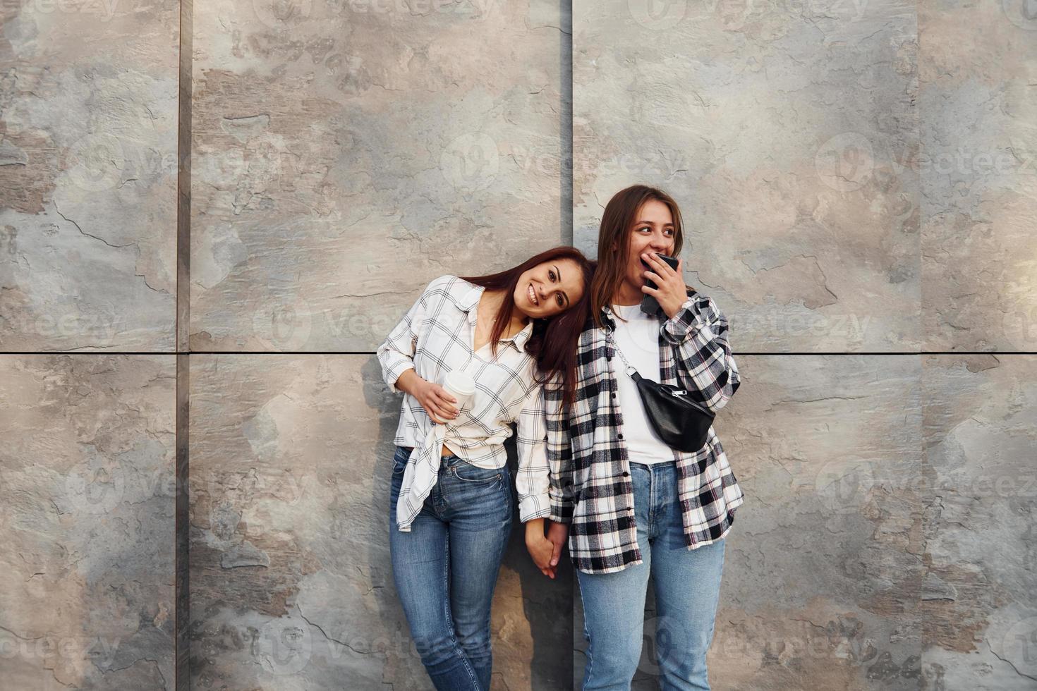 segurando a bebida e o telefone. lindas amigas alegres ou casal de lésbicas juntos perto da parede ao ar livre durante o dia foto