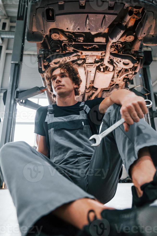 com ferramenta na mão. homem adulto em uniforme de cor cinza trabalha no salão do automóvel foto