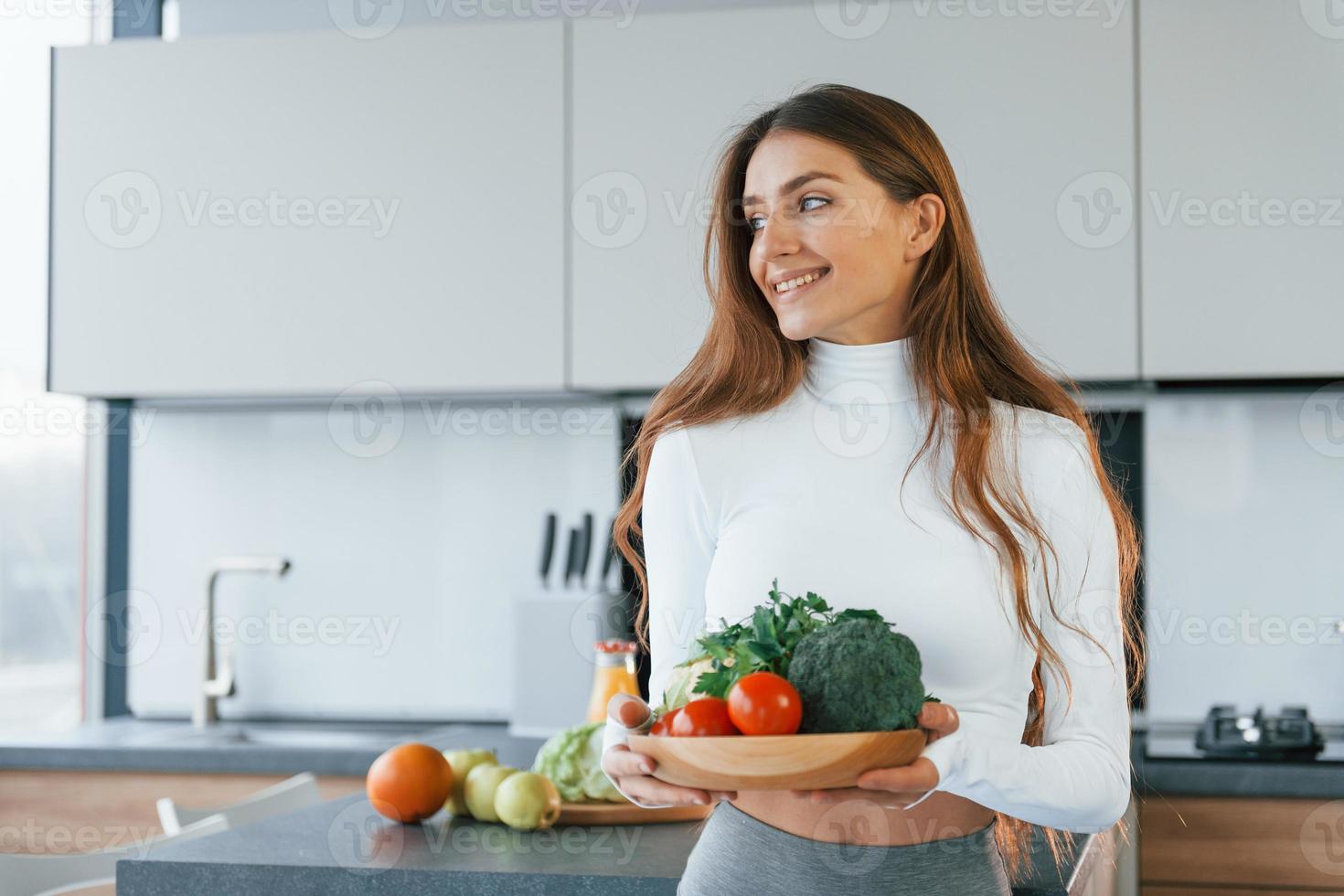 segura o prato com legumes. jovem europeia está dentro de casa na cozinha dentro de casa com comida saudável foto