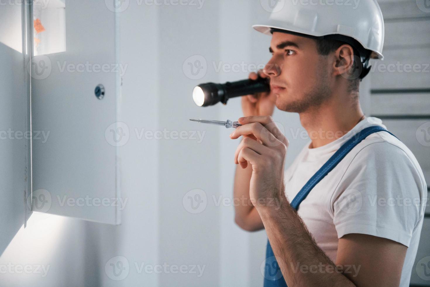 jovem eletricista masculino no capacete branco trabalha dentro de casa na sala foto