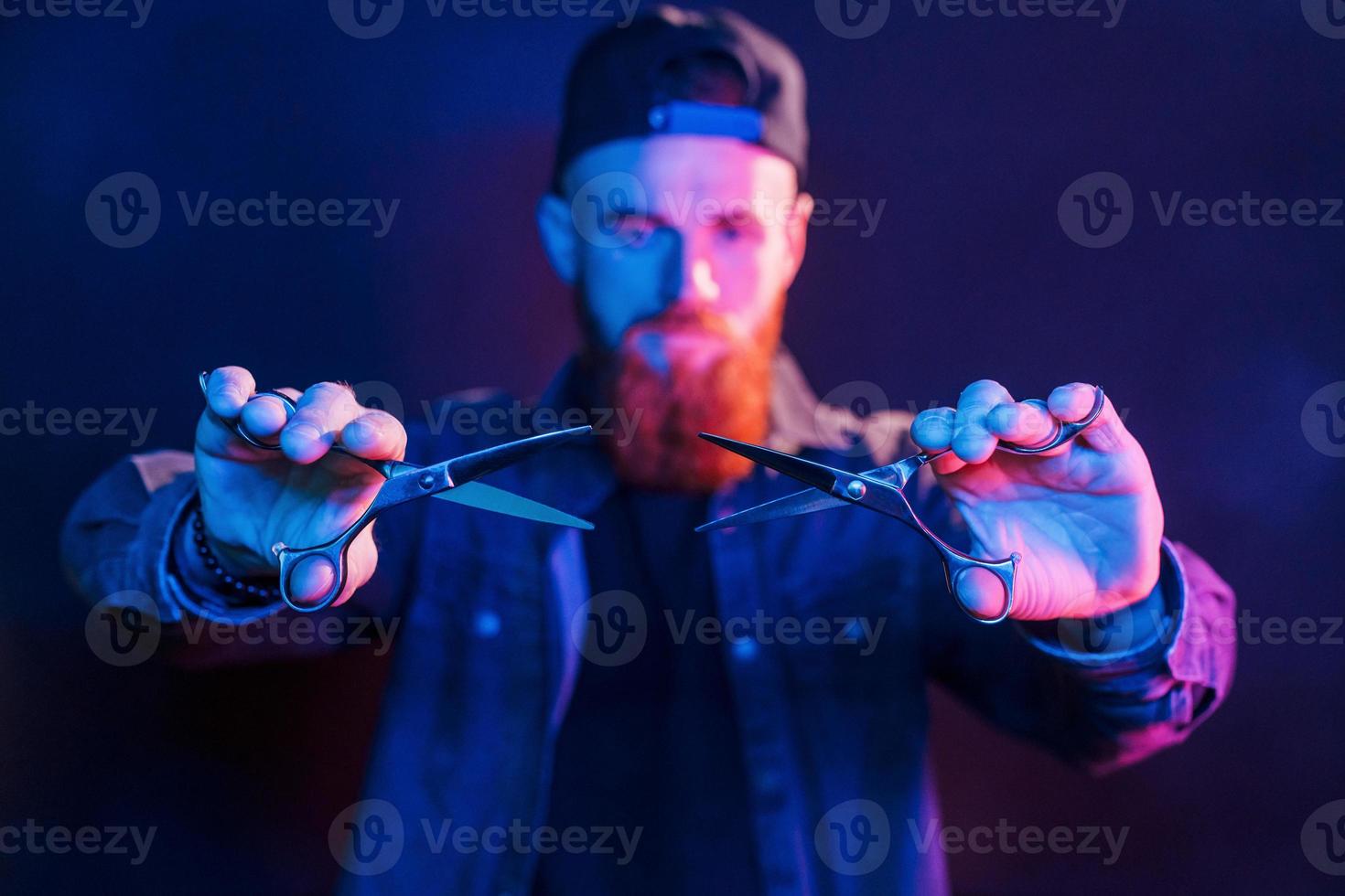 com tesoura. jovem barbeiro barbudo de boné parado no estúdio com iluminação neon foto