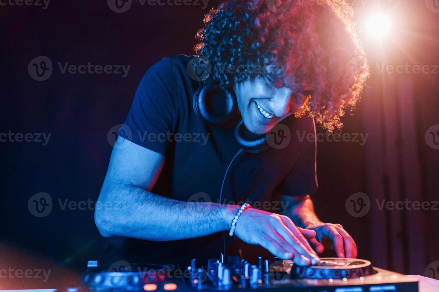 homem com cabelo encaracolado usando equipamento de dj e parado na sala escura iluminada por neon foto