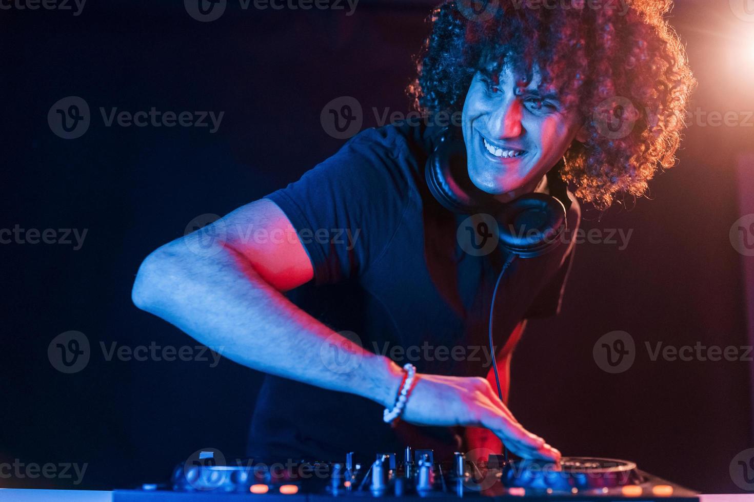 homem com cabelo encaracolado usando equipamento de dj e parado na sala escura iluminada por neon foto