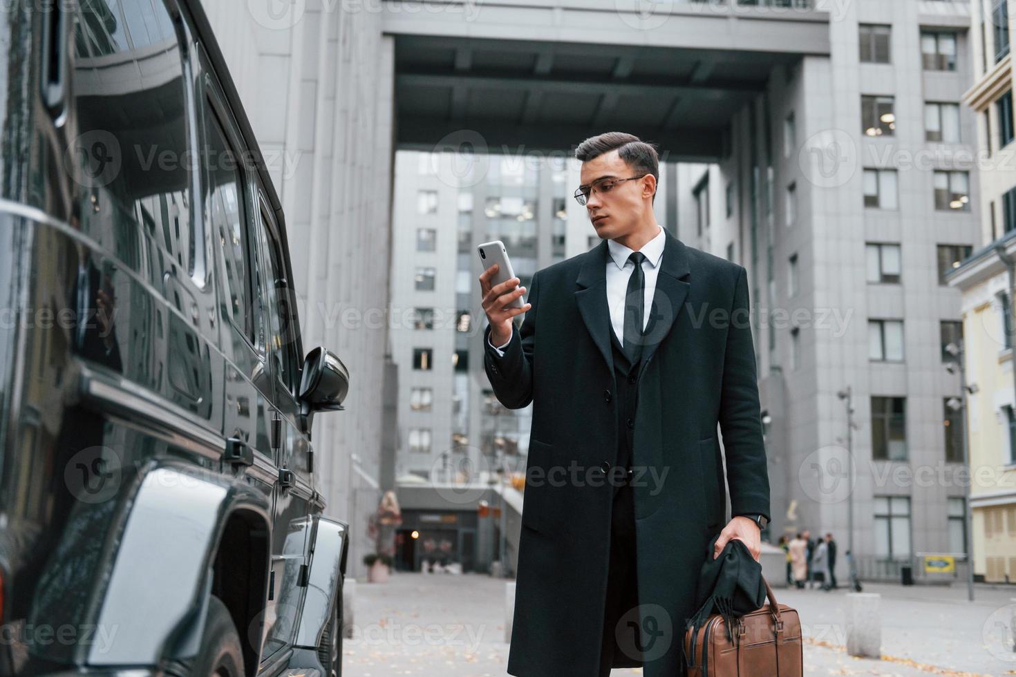 de pé perto de carro preto. empresário de terno preto e gravata está ao ar livre na cidade foto