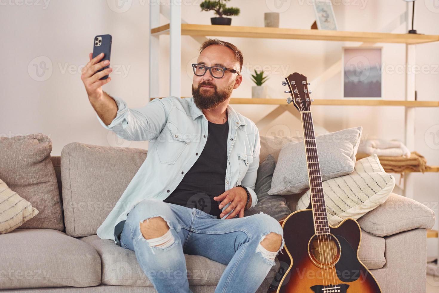sentado no sofá. homem em roupas casuais e com violão está dentro de casa foto