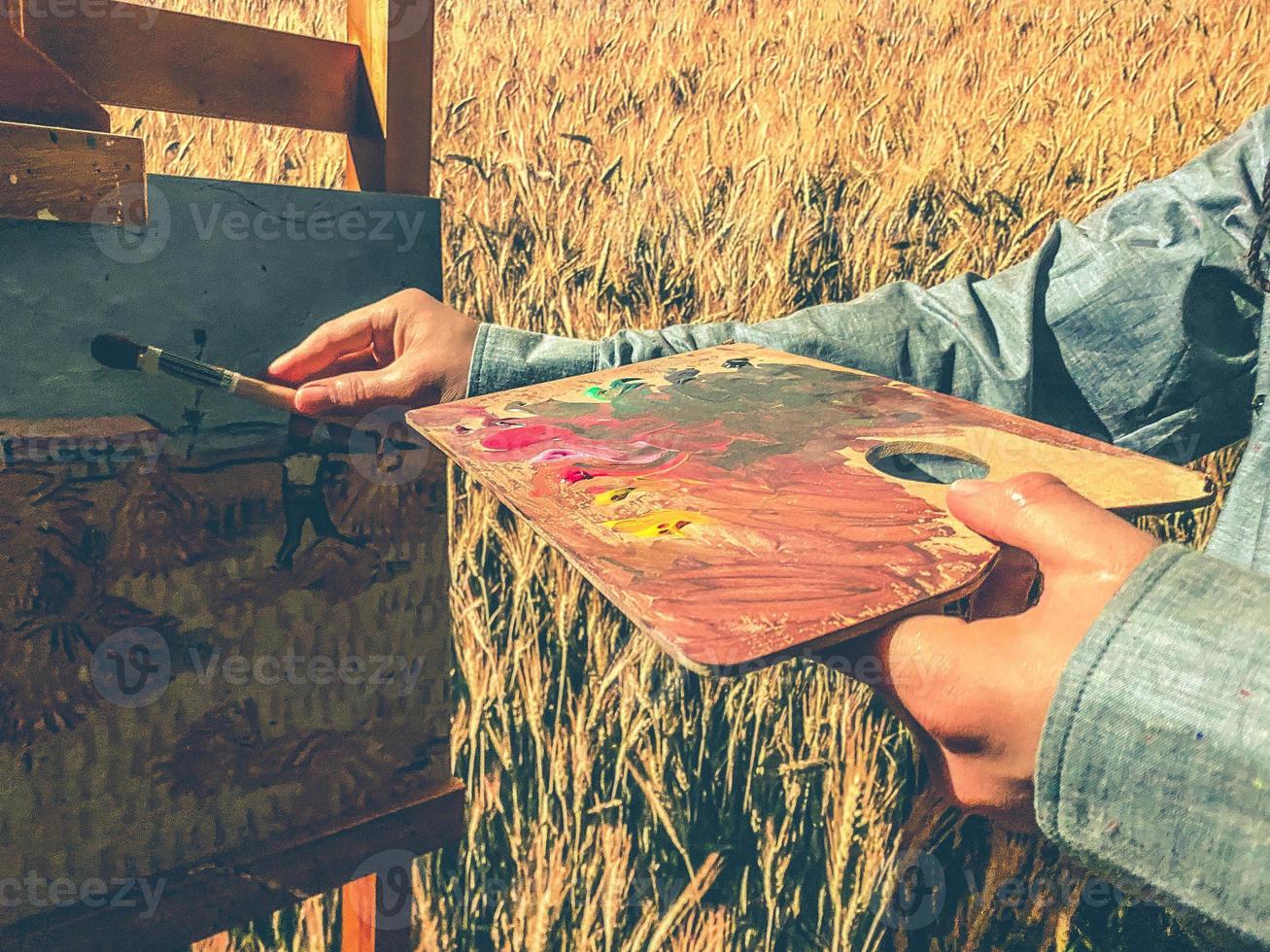 o artista pinta sobre tela. nas mãos do artista um pincel e uma paleta de tintas. misturou várias cores para pintar um quadro. homem desenha no fundo de um campo amarelo foto