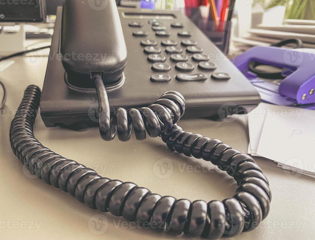 telefone fixo no escritório em cima da mesa. monofone preto com botões e display. meios de comunicação no escritório. fio preto no tubo foto