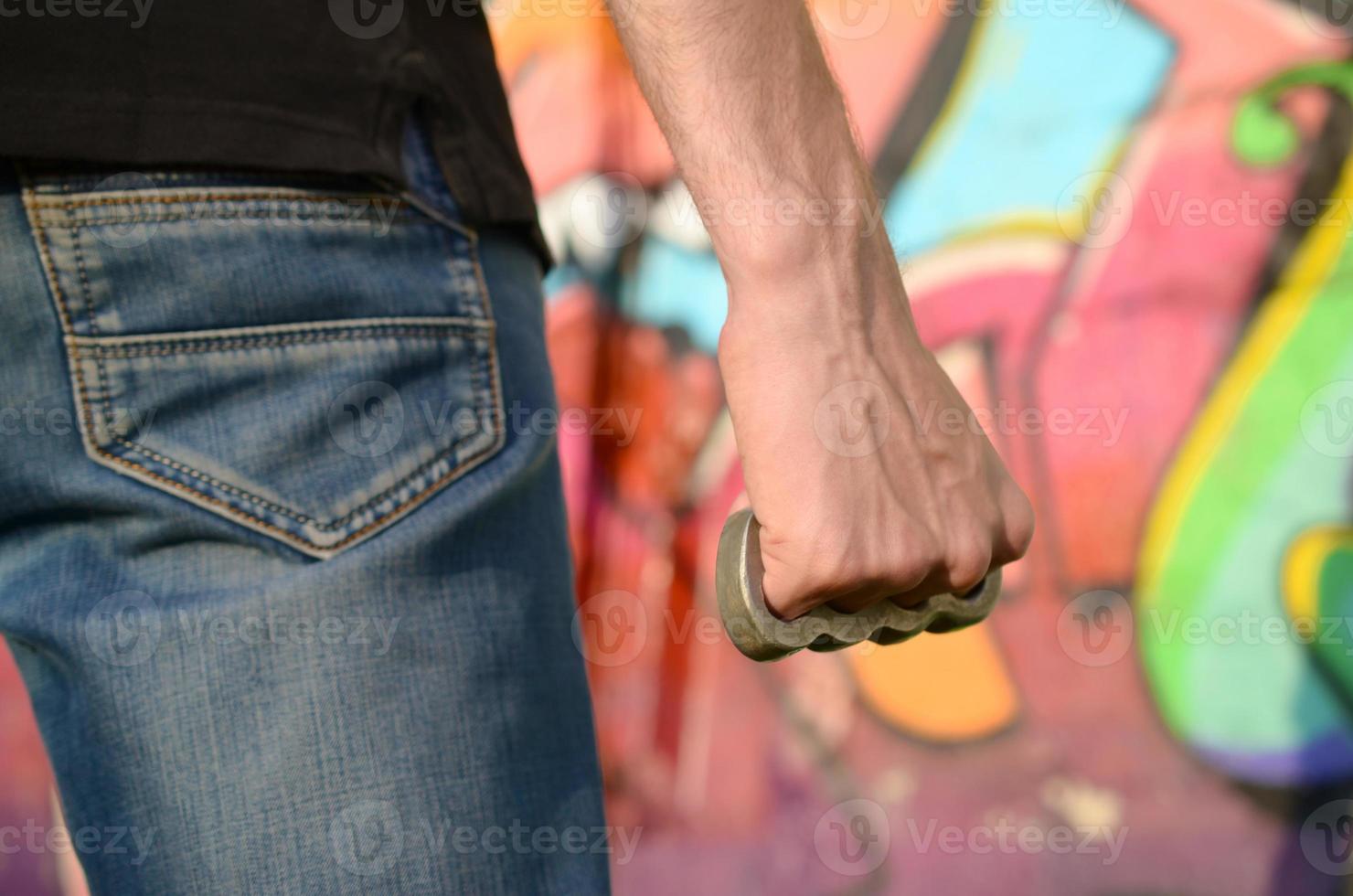 vista traseira do jovem homem caucasiano com junta de latão na mão contra a parede de tijolos do gueto com pinturas de grafite. conceito de forças criminosas e acusação de agressão foto