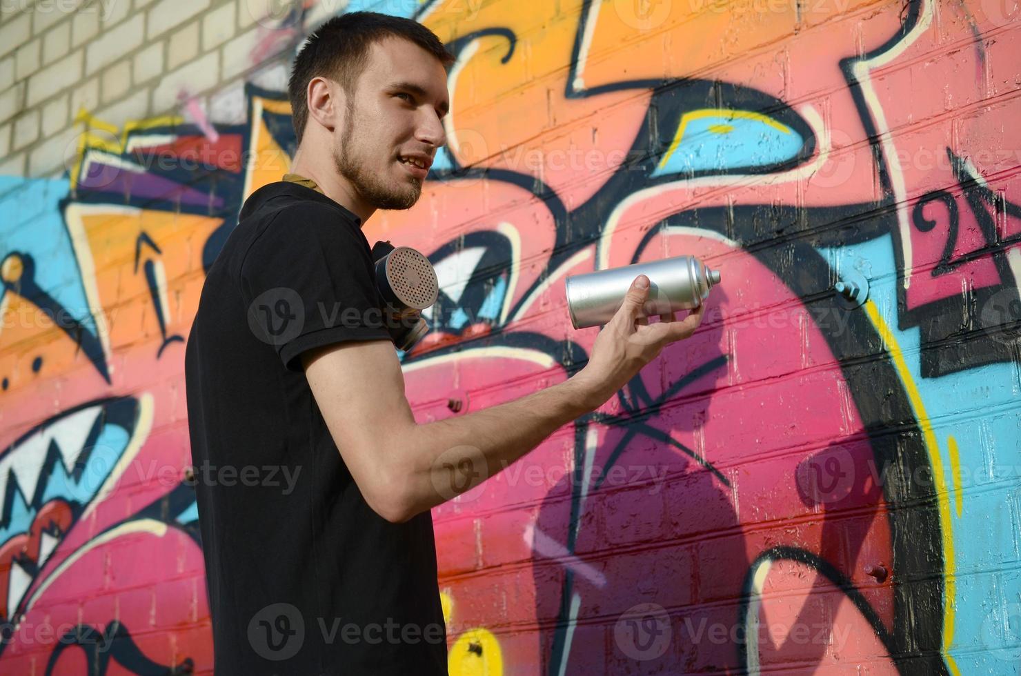 jovem grafiteiro com mochila e máscara de gás no pescoço pinta graffiti colorido em tons de rosa na parede de tijolos. arte de rua e processo de pintura contemporânea foto