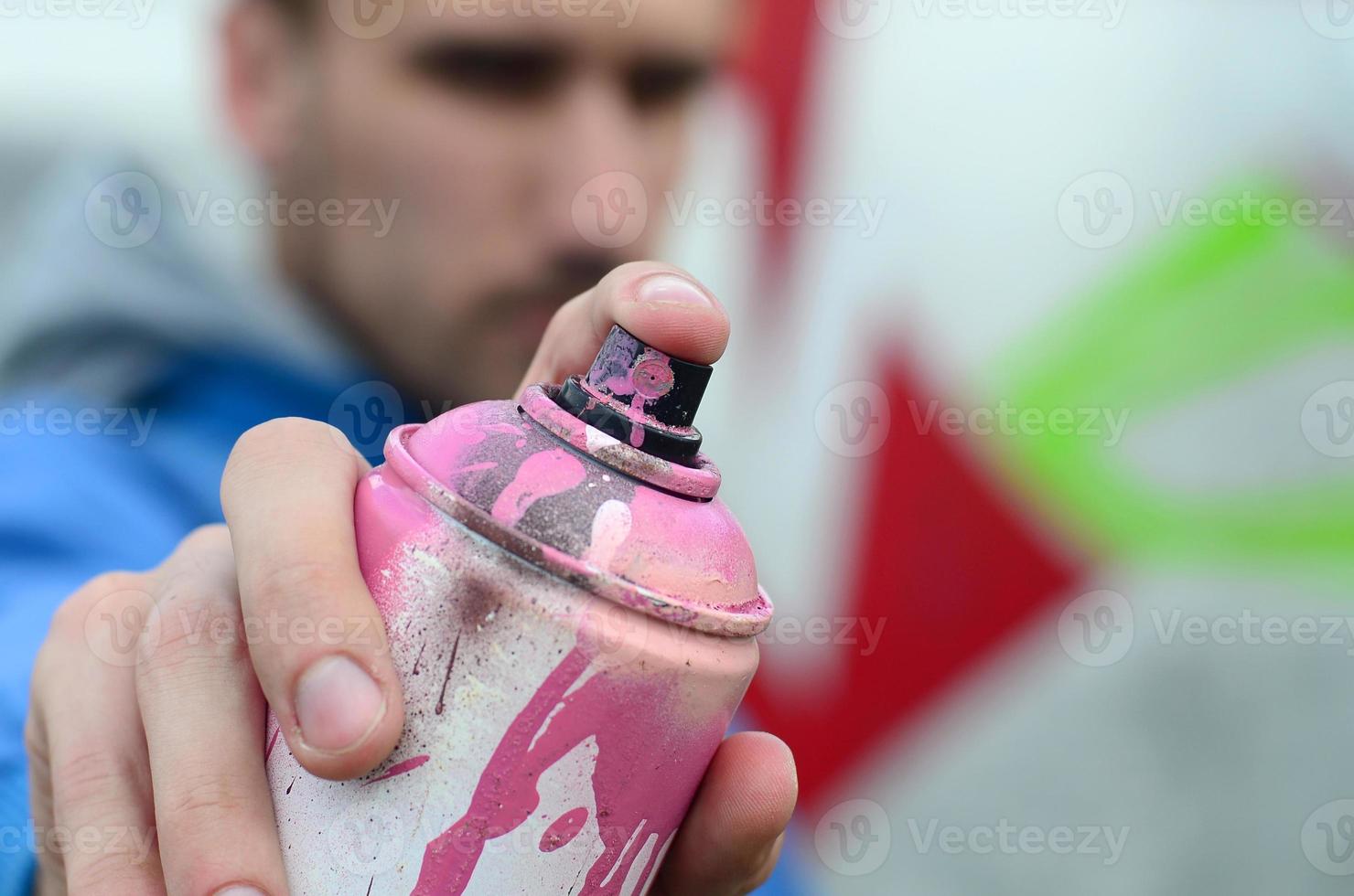 um jovem grafiteiro em uma jaqueta azul está segurando uma lata de tinta na frente dele contra um fundo de desenho de grafite colorido. arte de rua e conceito de vandalismo foto