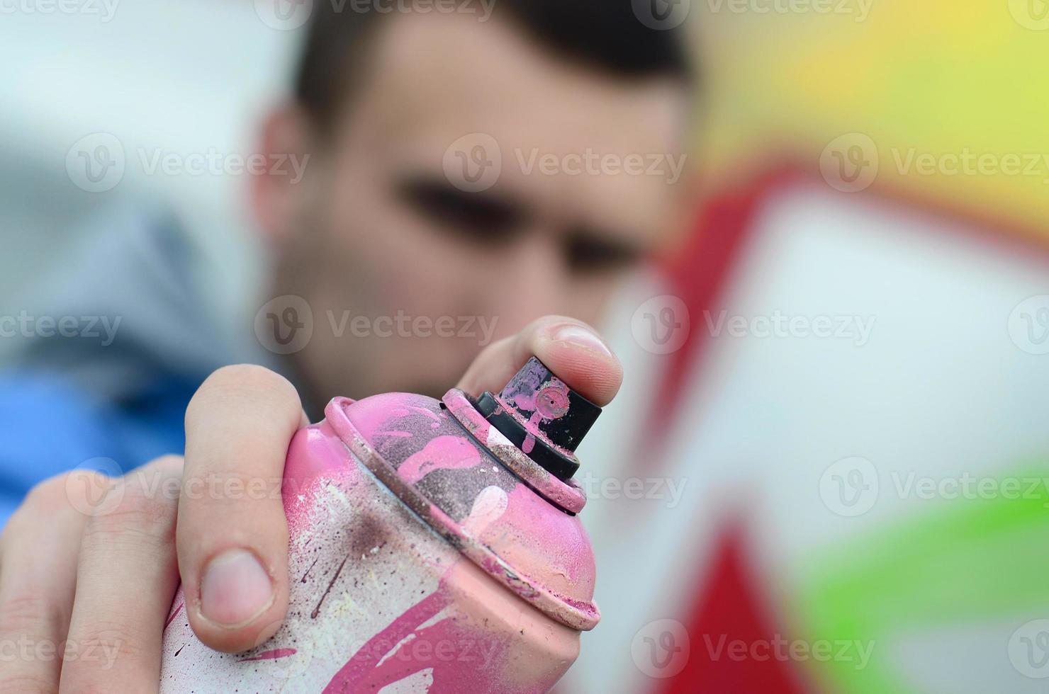 um jovem grafiteiro em uma jaqueta azul está segurando uma lata de tinta na frente dele contra um fundo de desenho de grafite colorido. arte de rua e conceito de vandalismo foto