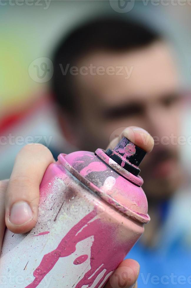 um jovem grafiteiro em uma jaqueta azul está segurando uma lata de tinta na frente dele contra um fundo de desenho de grafite colorido. arte de rua e conceito de vandalismo foto