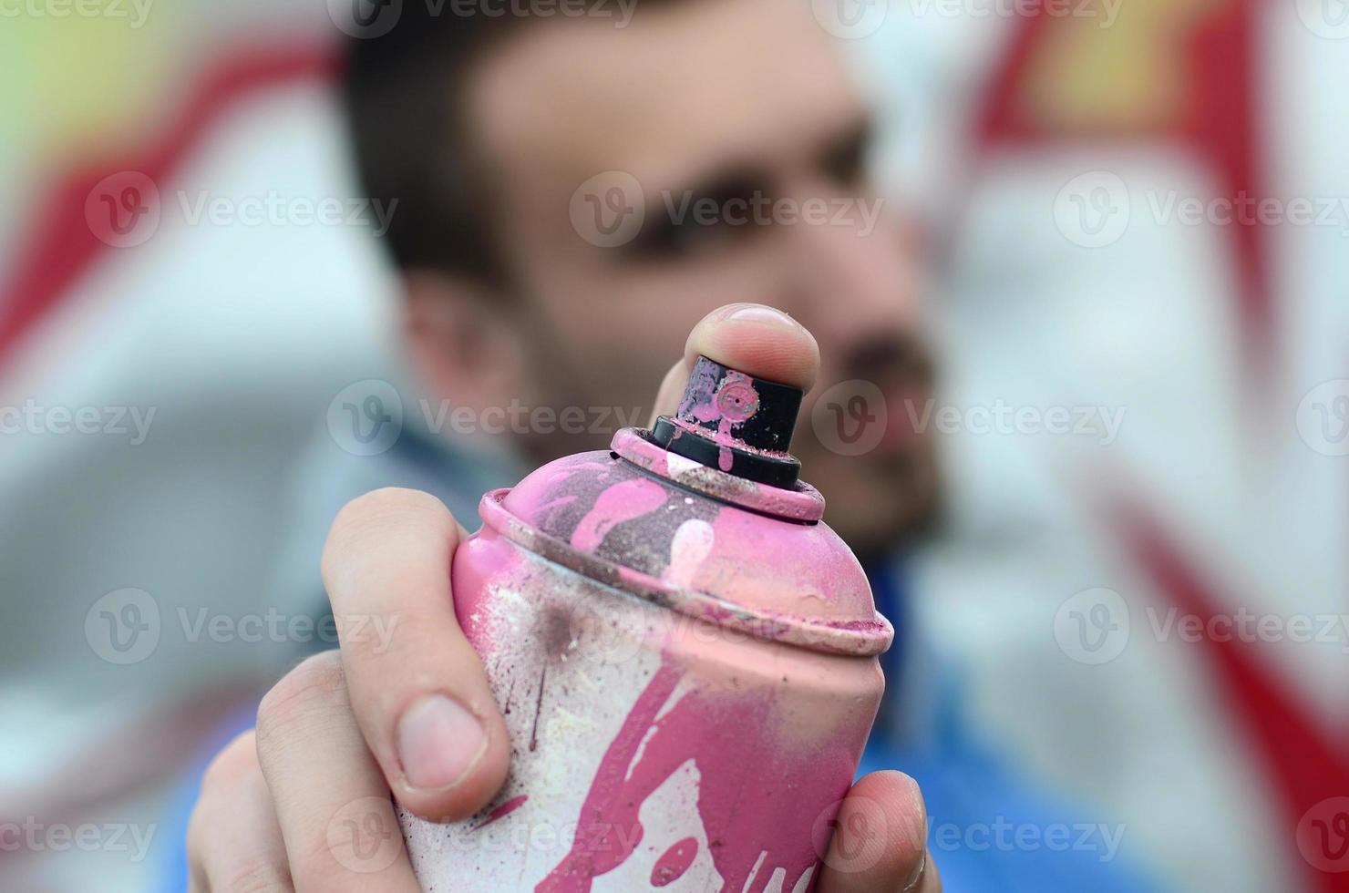 um jovem grafiteiro em uma jaqueta azul está segurando uma lata de tinta na frente dele contra um fundo de desenho de grafite colorido. arte de rua e conceito de vandalismo foto