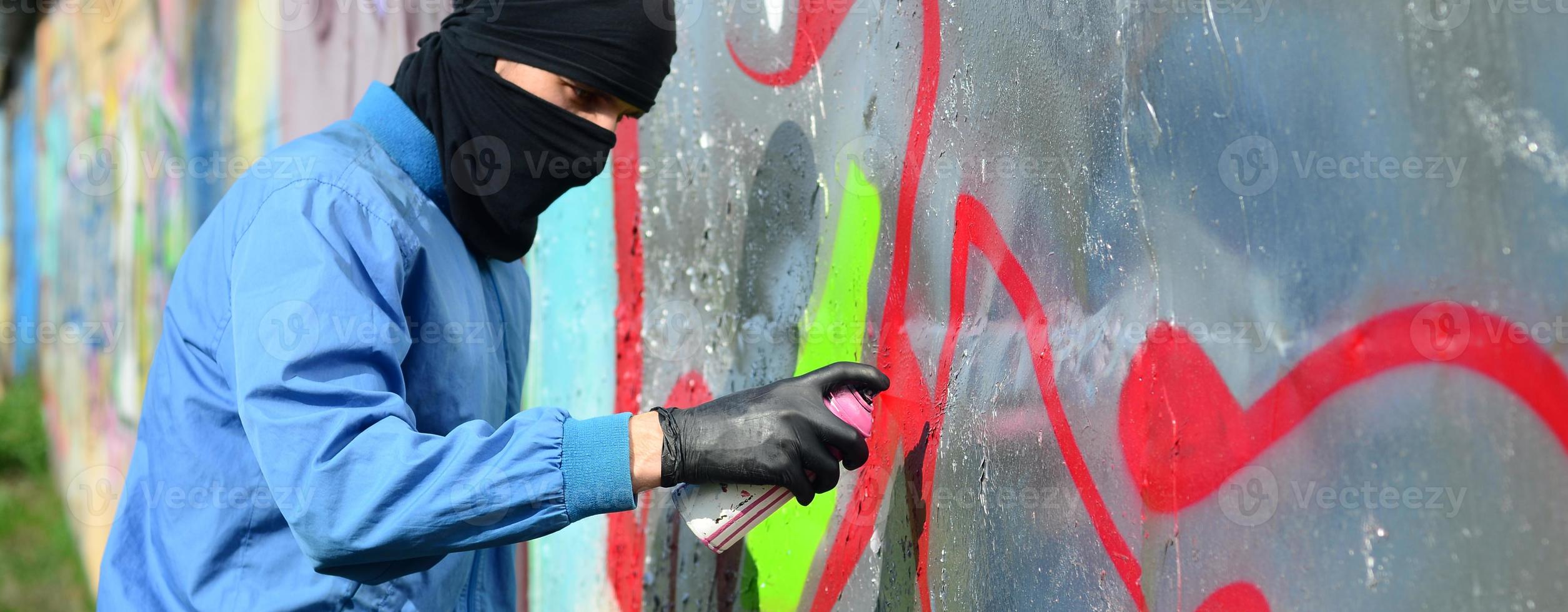 um jovem hooligan com um rosto escondido pinta graffiti em uma parede de metal. conceito de vandalismo ilegal foto