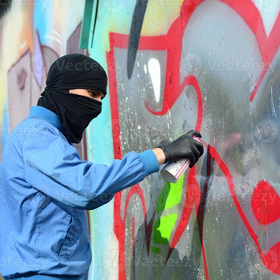 um jovem hooligan com um rosto escondido pinta graffiti em uma parede de metal. conceito de vandalismo ilegal foto