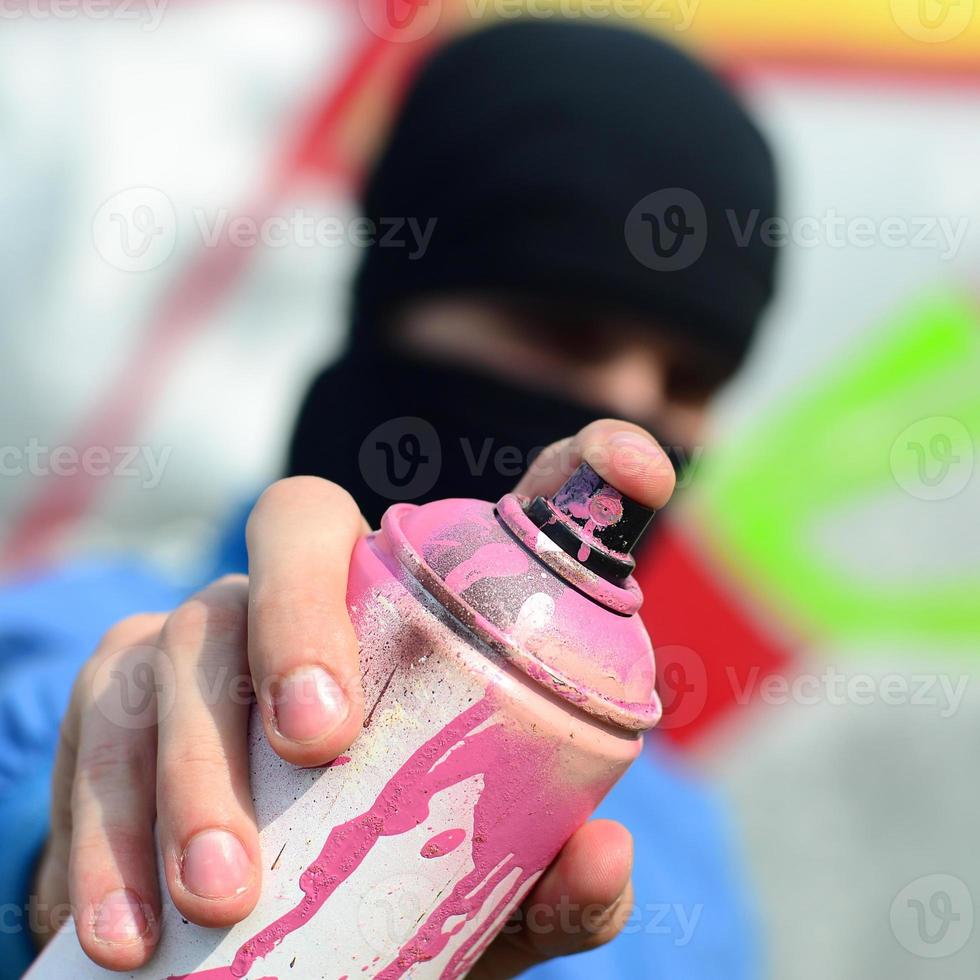 um jovem grafiteiro em uma jaqueta azul e máscara preta está segurando uma lata de tinta na frente dele contra um fundo de desenho de graffiti colorido. arte de rua e conceito de vandalismo foto