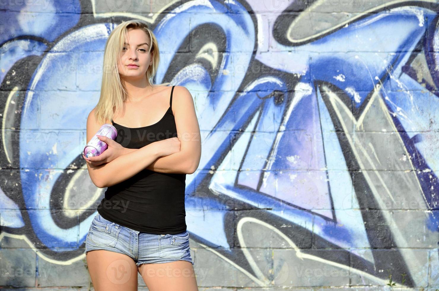 uma jovem e bela garota sexy grafiteira com um spray de tinta fica no fundo da parede com um padrão de graffiti em tons de azul e roxo foto