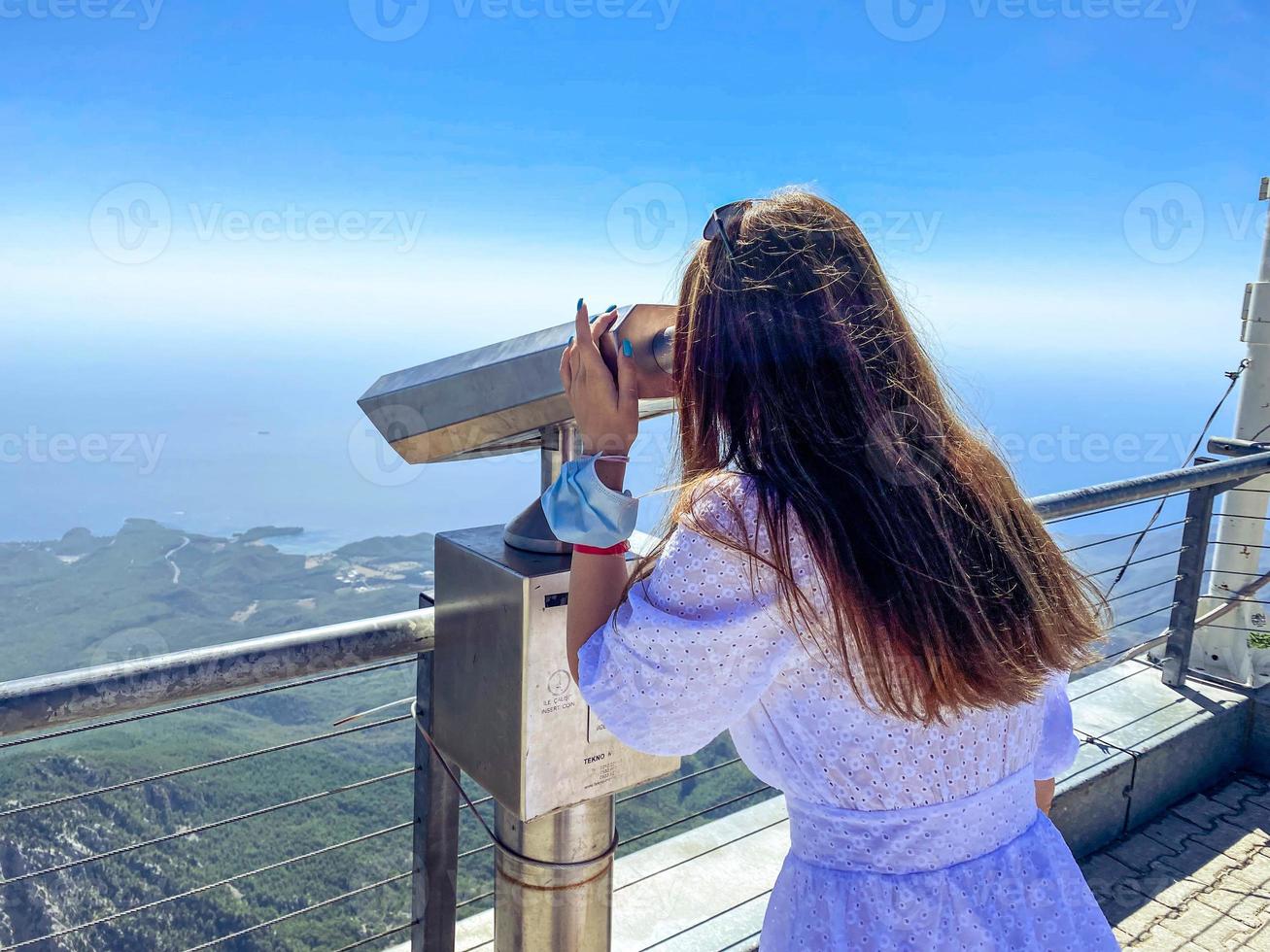 uma garota de vestido branco com cabelo comprido olha através de binóculos em uma plataforma de observação. passeio turístico, turista curioso foto