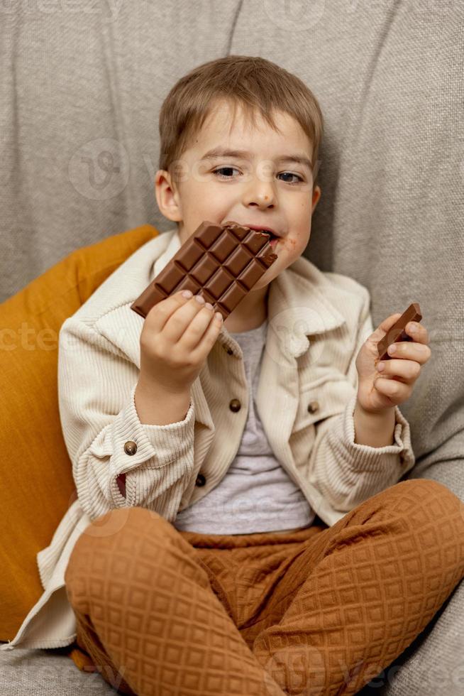 menino adorável sentado no sofá em casa e comendo barra de chocolate. criança e doces, confeitos de açúcar. criança desfrutar de uma deliciosa sobremesa. criança pré-escolar com roupas casuais. emoção positiva. foto