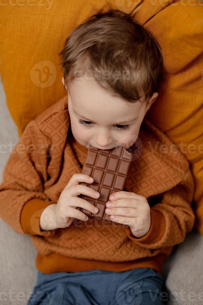 menino adorável sentado no sofá em casa e comendo barra de chocolate. criança e doces, confeitos de açúcar. criança desfrutar de uma deliciosa sobremesa. criança pré-escolar com roupas casuais. emoção positiva. foto