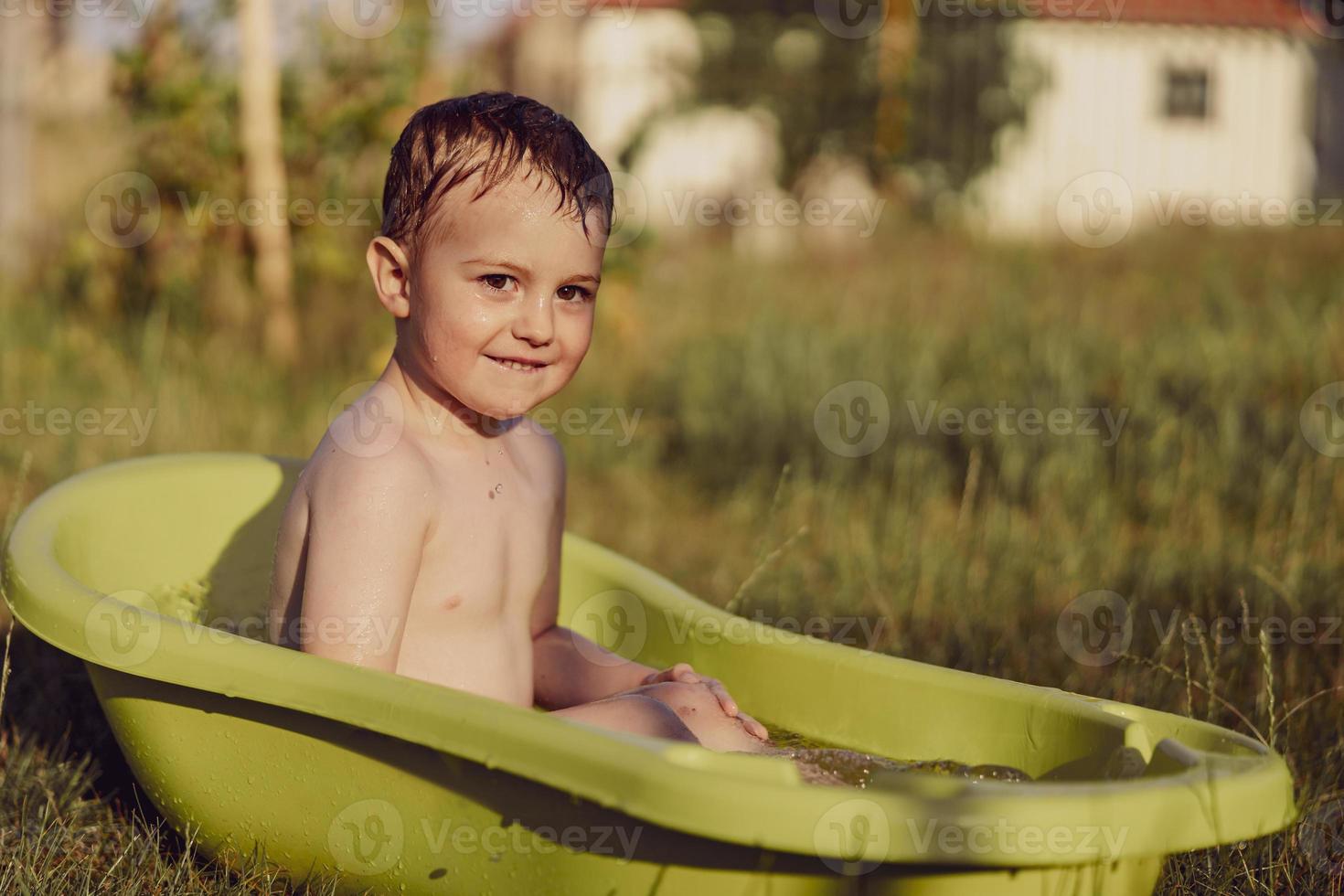 menino bonitinho tomando banho na banheira ao ar livre no jardim. criança feliz está espirrando, brincando com água e se divertindo. temporada de verão e recreação. ficar fresco no calor do verão. diversão aquática no quintal. foto