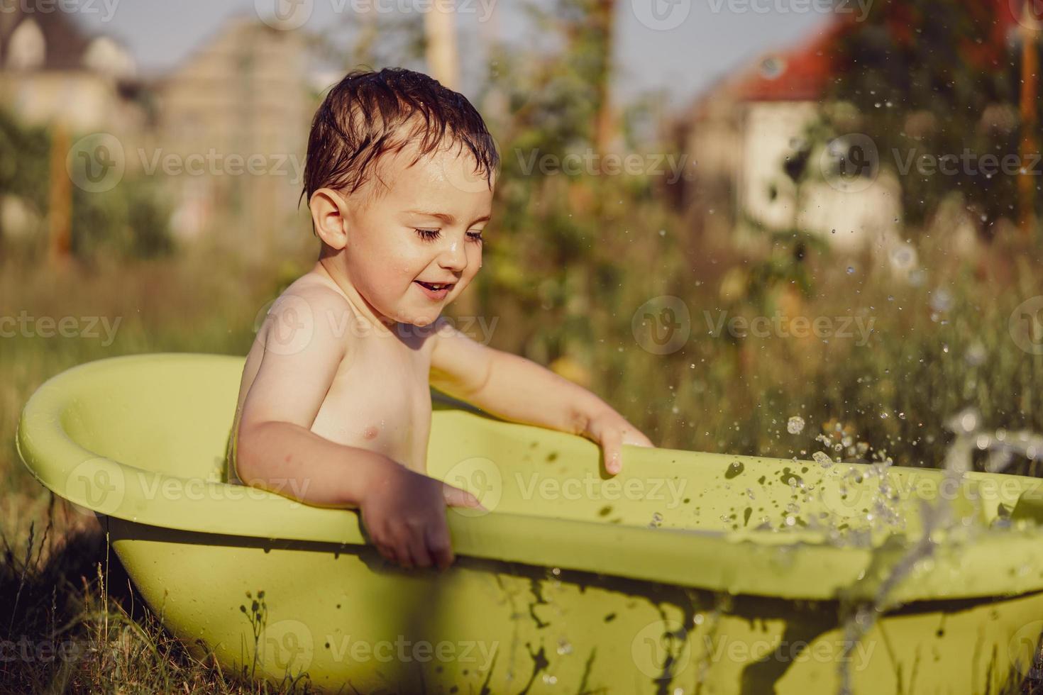 menino bonitinho tomando banho na banheira ao ar livre no jardim. criança feliz está espirrando, brincando com água e se divertindo. temporada de verão e recreação. ficar fresco no calor do verão. diversão aquática no quintal. foto