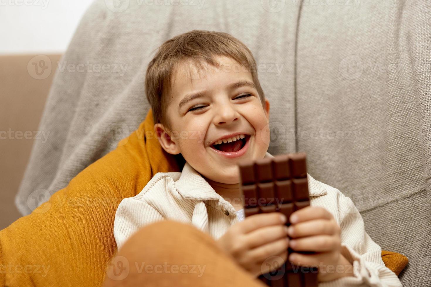 menino adorável sentado no sofá em casa e comendo barra de chocolate. criança e doces, confeitos de açúcar. criança desfrutar de uma deliciosa sobremesa. criança pré-escolar com roupas casuais. emoção positiva. foto