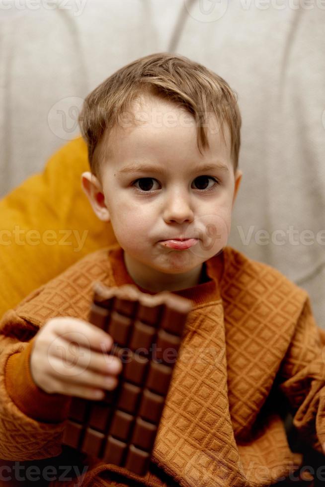 menino adorável sentado no sofá em casa e comendo barra de chocolate. criança e doces, confeitos de açúcar. criança desfrutar de uma deliciosa sobremesa. criança pré-escolar com roupas casuais. foto