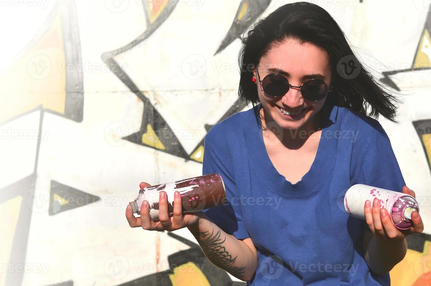 retrato de uma jovem emocional com cabelo preto e piercings. foto de uma menina com latas de tinta aerossol nas mãos em um fundo de parede de graffiti. o conceito de arte de rua e uso de tintas aerossóis