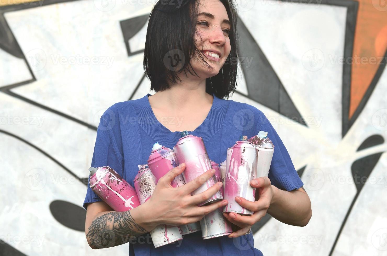 retrato de uma jovem emocional com cabelo preto e piercings. foto de uma menina com latas de tinta aerossol nas mãos em um fundo de parede de graffiti. o conceito de arte de rua e uso de tintas aerossóis