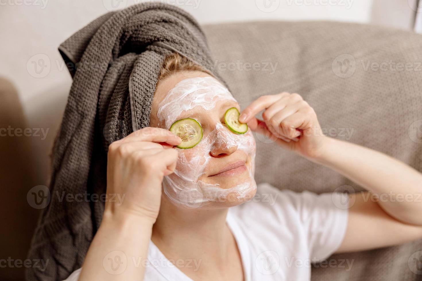 jovem mulher com roupas casuais, toalha na cabeça e máscara de beleza no rosto, sentada no sofá e descansando. relaxar depois de tomar banho. cosmético para mulheres, cuidados com a pele. autocuidado, tempo para você. relaxante, foto