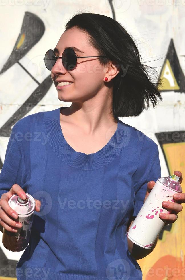 retrato de uma jovem emocional com cabelo preto e piercings. foto de uma menina com latas de tinta aerossol nas mãos em um fundo de parede de graffiti. o conceito de arte de rua e uso de tintas aerossóis