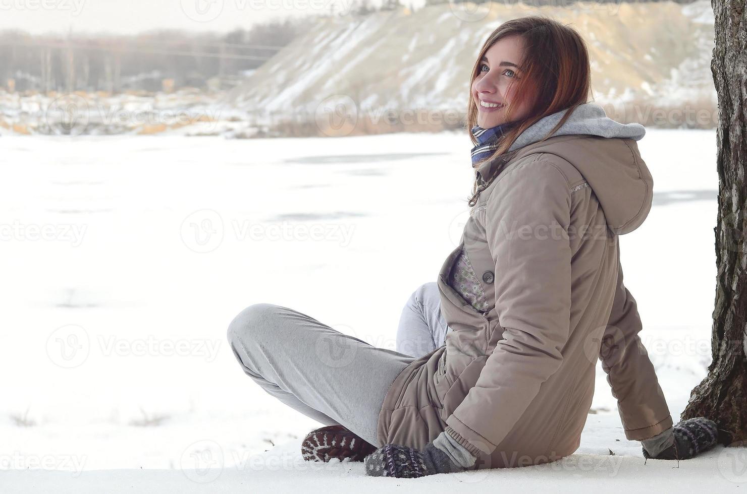 uma jovem caucasiana com um casaco marrom está sentada perto de um penhasco no fundo de uma linha do horizonte entre o céu e um lago congelado no inverno foto