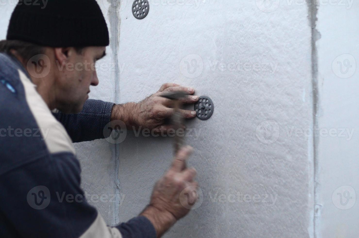 um trabalhador idoso enfia um pino em um guarda-chuva de plástico em uma parede de isopor. o processo de fixação de placas de poliestireno expandido. aquecimento da fachada do prédio foto