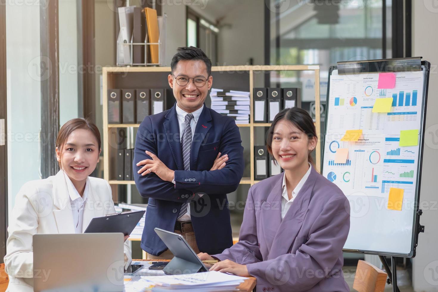 equipe de negócios três homens e mulheres asiáticos sorridentes e felizes, usando tablets e laptops, debatem e traçam estatísticas em um quadro de gráficos corporativos dentro da empresa. conceito de colegas. foto