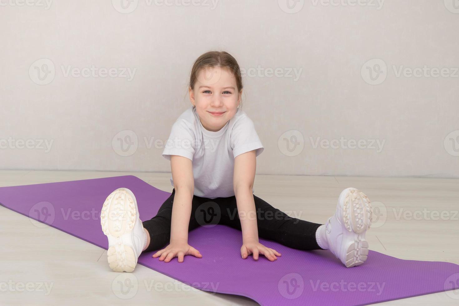 menina bonita em um uniforme esportivo senta-se em uma esteira de ginástica com os braços estendidos para a frente e sorri foto