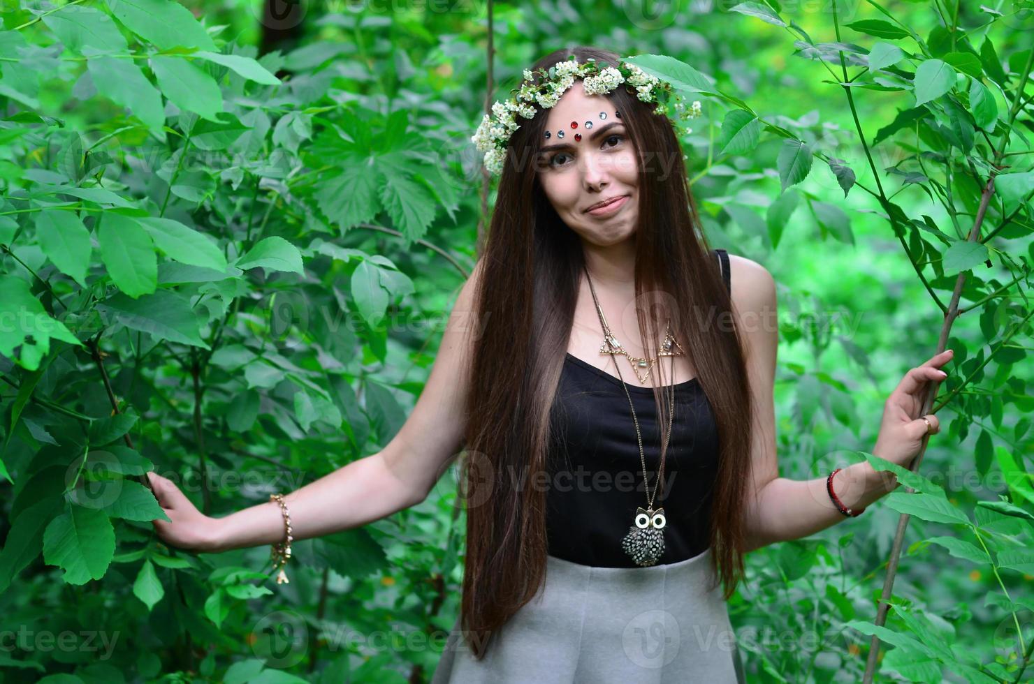 uma foto da floresta de uma bela jovem morena de aparência européia com olhos castanhos escuros e lábios grandes. na cabeça da menina está uma coroa de flores, na testa enfeites brilhantes