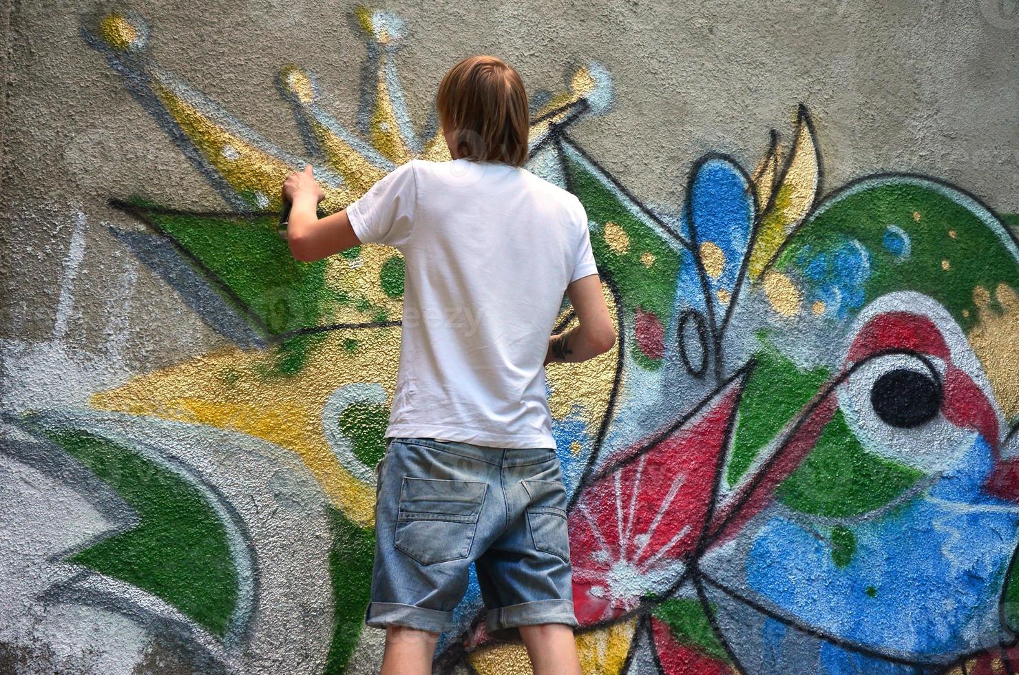foto de um jovem de short jeans e camisa branca. o cara desenha na parede do grafite um desenho com tintas aerossol de várias cores. o conceito de hooliganismo e danos à propriedade