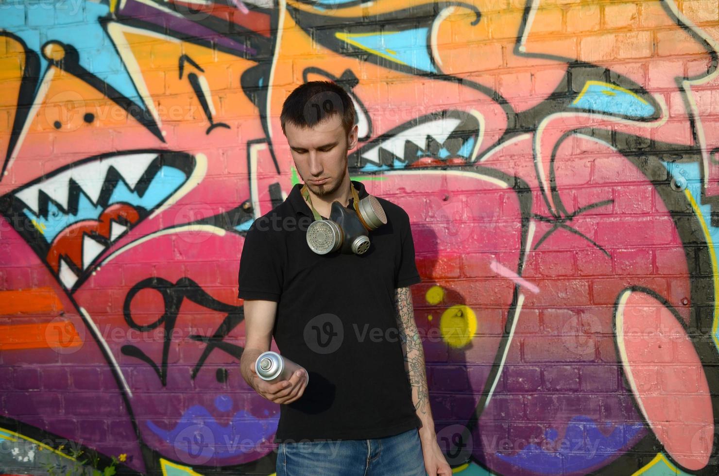 jovem grafiteiro caucasiano em camiseta preta com spray de aerossol prata pode perto de graffiti colorido em tons de rosa na parede de tijolos. arte de rua e processo de pintura contemporânea foto