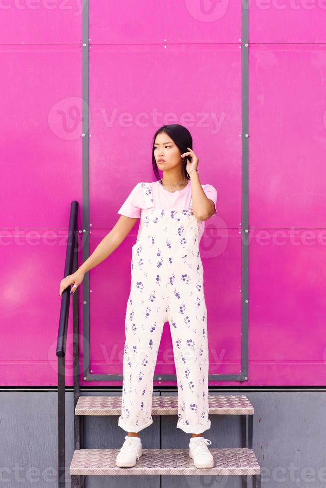 mulher chinesa com olhar vazio e expressão séria em pé contra a parede rosa do edifício moderno. foto